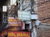 a sign in faded green reading "Discourage the Beggars/Chibahal Pariwar, Thamel"; surrounding it are multiple red signs with yellow writing for a music store, and both signs are on posts that feature a tangle of wires