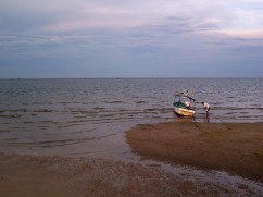 a Thai man is seen crouching next to his small boat on the beach; except for in the distance, there are no other boats and the sun has begun to set